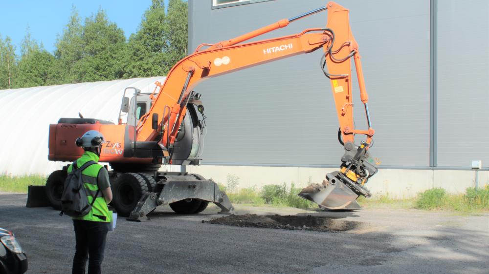 Kaivinkone kaivamassa kuoppaa, jota henkilö seuraa turvaliivi ja kypärä yllään.
