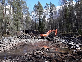 Kuva Äänekosken Mämmenkosken työmaasta. Kuvassa koski, jonka keskellä on oranssi kaivuri, kiviä, puita ja taivas.