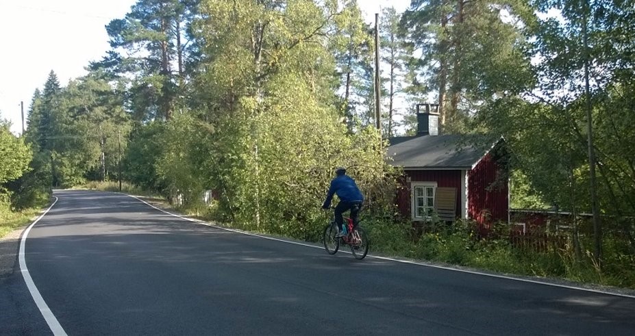 Mies pyöräilemässä punaisen puutalon ohitse asfalttipäällysteisellä tiellä. 