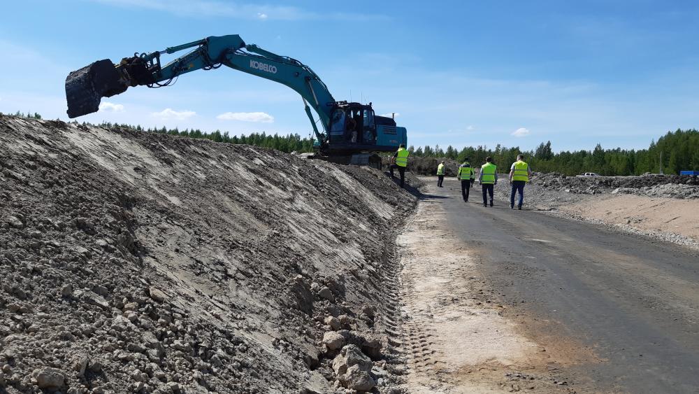 Etualalla kohonneen maanpinnan reunaa, taaempana kaivinkone ja ihmisiä huomioliiveissä.