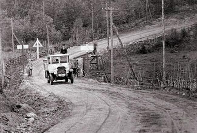Auto ajaa tiellä 100 vuotta sitten, tien mutkassa myös ensimmäinen liikennemerkki.
