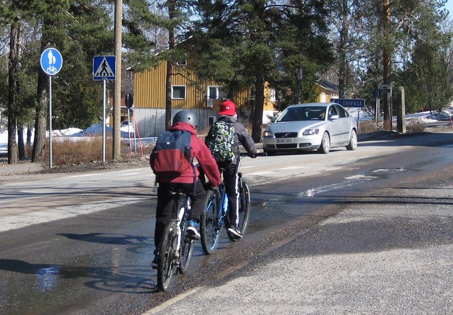 Kaksi nuorta polkevat oikeassa tienlaidassa taajamassa. Harmaa henkilöauto kohtaa heidät vastaantulevien kaistalla. 