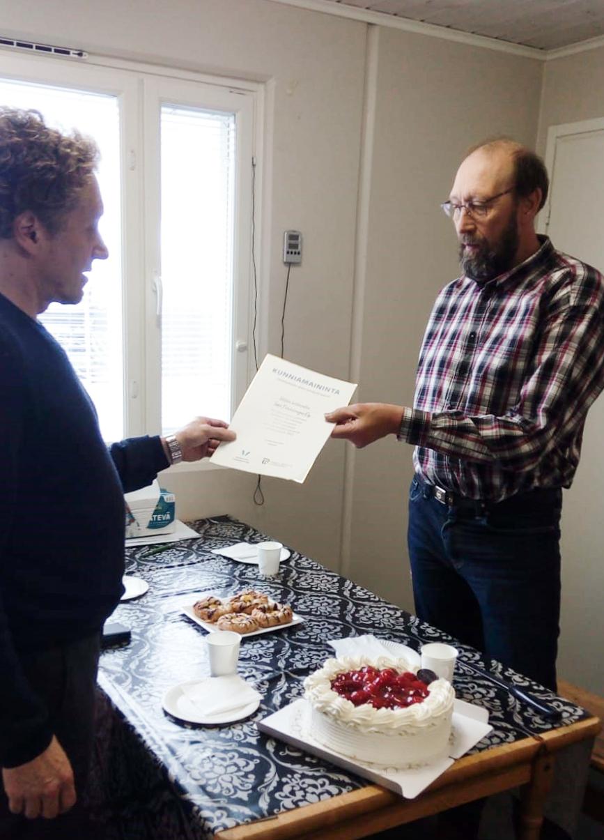 Aluevastaava Mikael Roiha ojentaa kunniakirjan yrittäjä Tapio Pahkakankaalle kakkukahvipöydän ääressä.