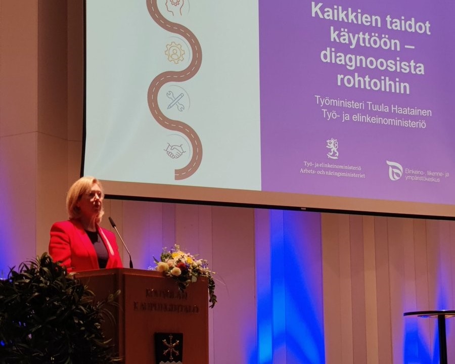 Ministeri Haatainen puhumassa puhujapöntössä, taustalla diaesitys missä tapahtuman nimi.