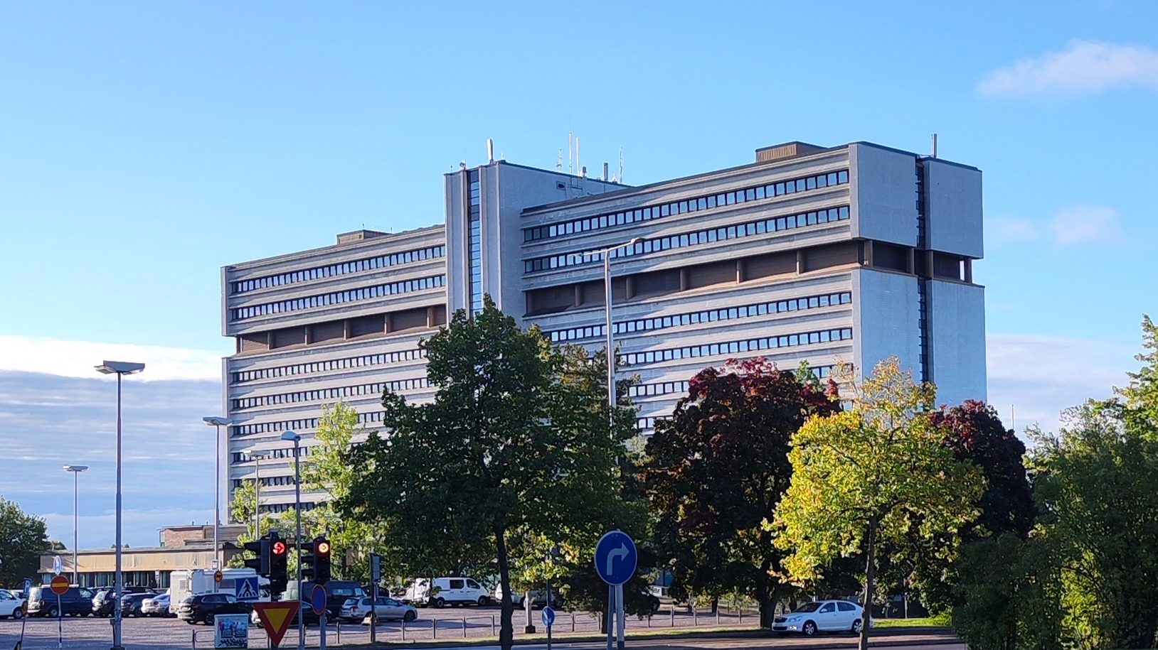 Suuri valkoinen rakennus, edustalla autoja, sininen taivas.