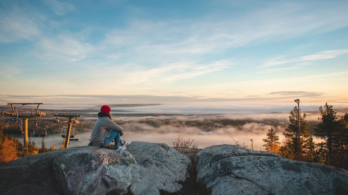 Punapipoinen henkilö istuu korkealla kalliolla hiihtohissien vierellä katsoen sumuiseen maisemaan.