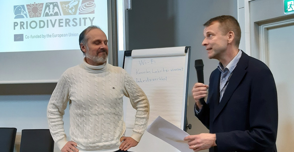 Kaksi miestä seisomassa valkokankaan edessä; toisella mikrofoni kädessä. 