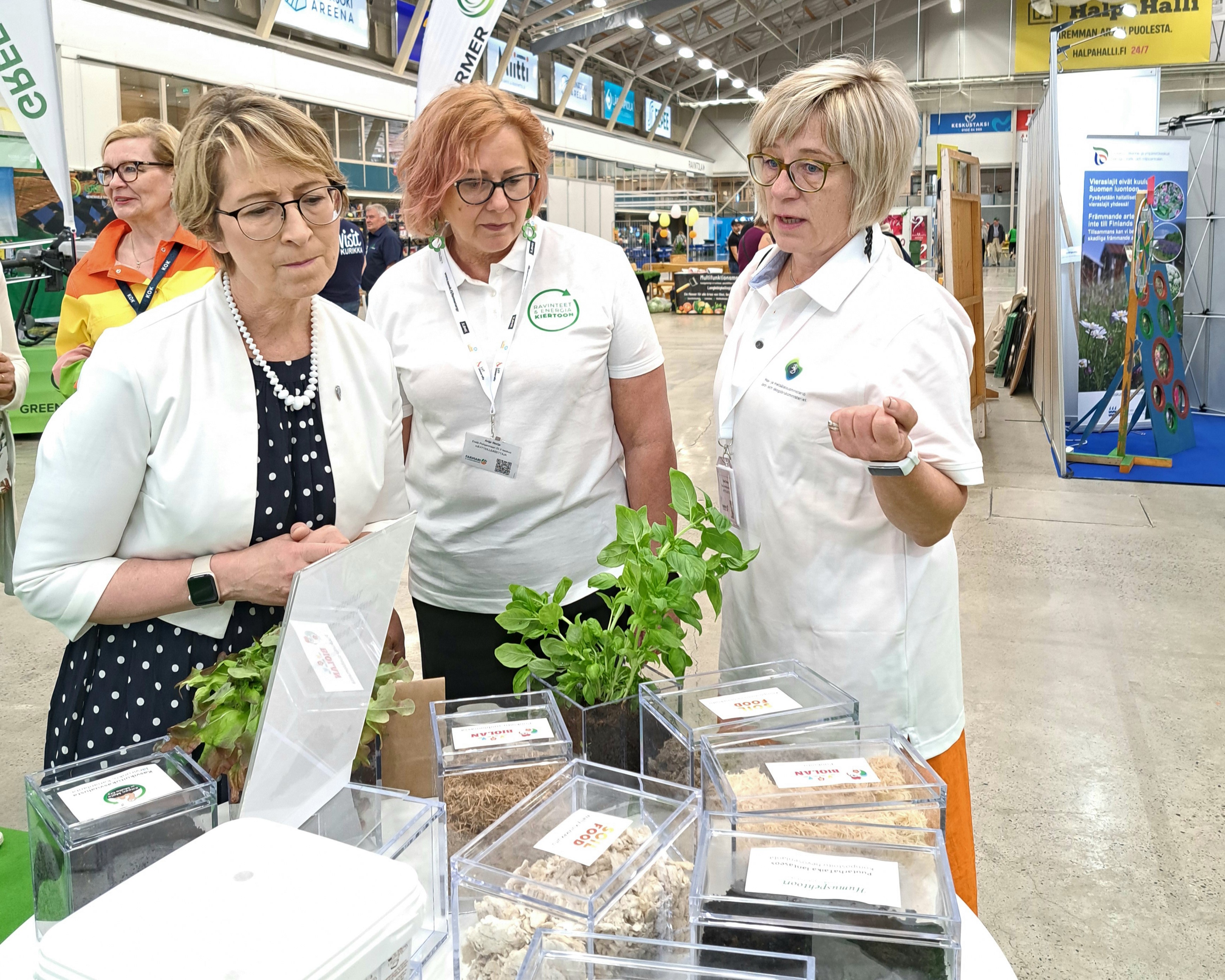 Kuvassa Anja Norja ja Sanna Tikander esittelevät maa- ja metsätalousministeri Sari Essayahille kierrätysravinteita Farmari-messuilla. Kolmikko seisoo näytepöydän edessä.
