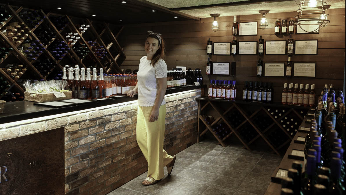 Nainen seisoo myyntitiskillä. Pöydillä ja seinillä on paljon viinipulloja aseteltu esille.