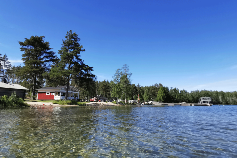 Järvenrantamaisema järveltä päin kuvattuna. Hiekkaranta, mökkejä, puita ja iso laituri