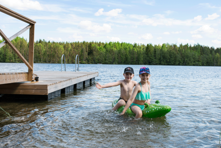 Tyttö ja poika istuvat puhallettavan vesilelun päällä järvivedessä. Taustalla laituri ja järvimaisema.