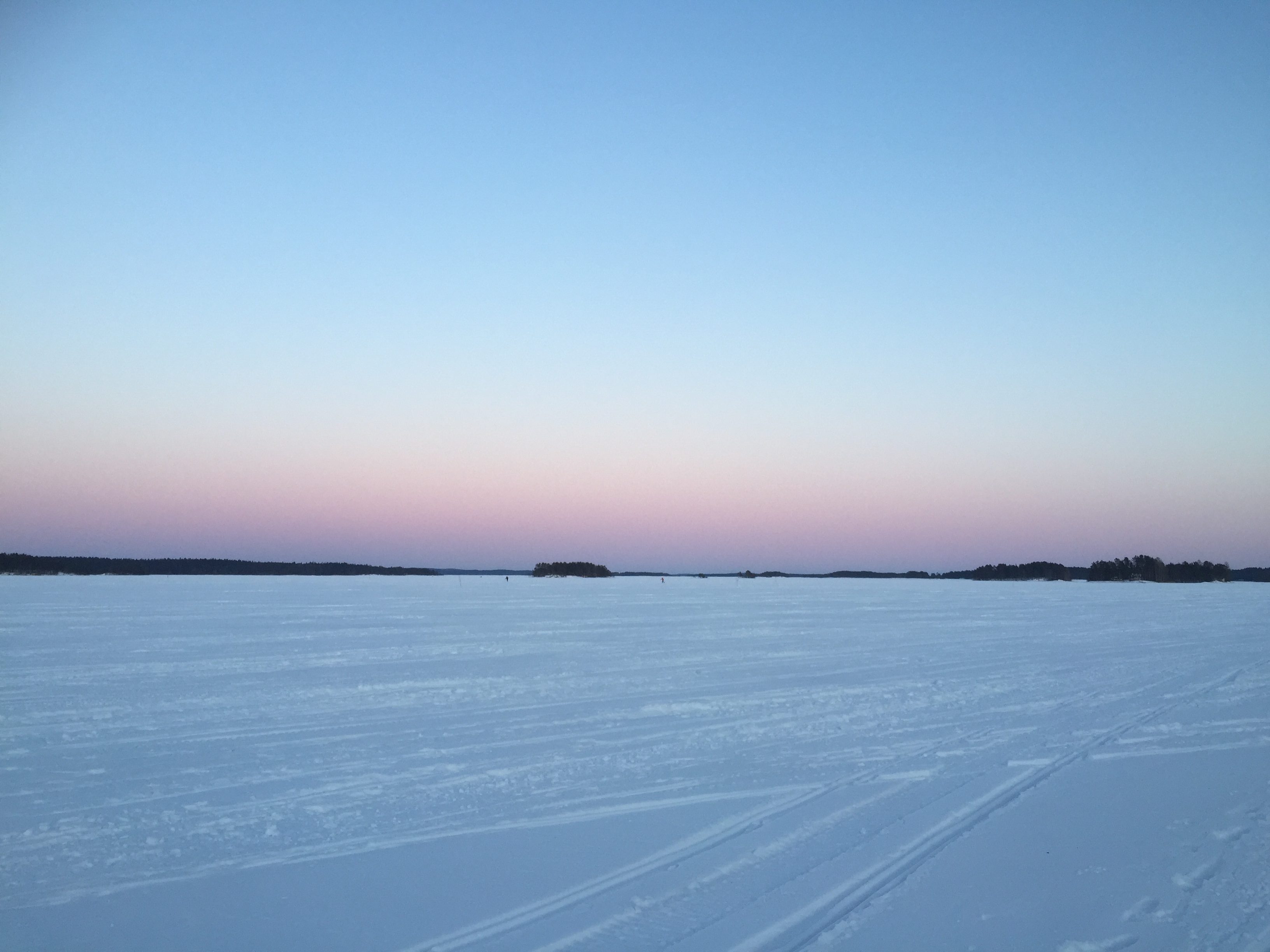 Kuva talviselta järven jäältä, jossa auringonlaskun viimeiset säteet näkyvät horisontissa puiden takaa