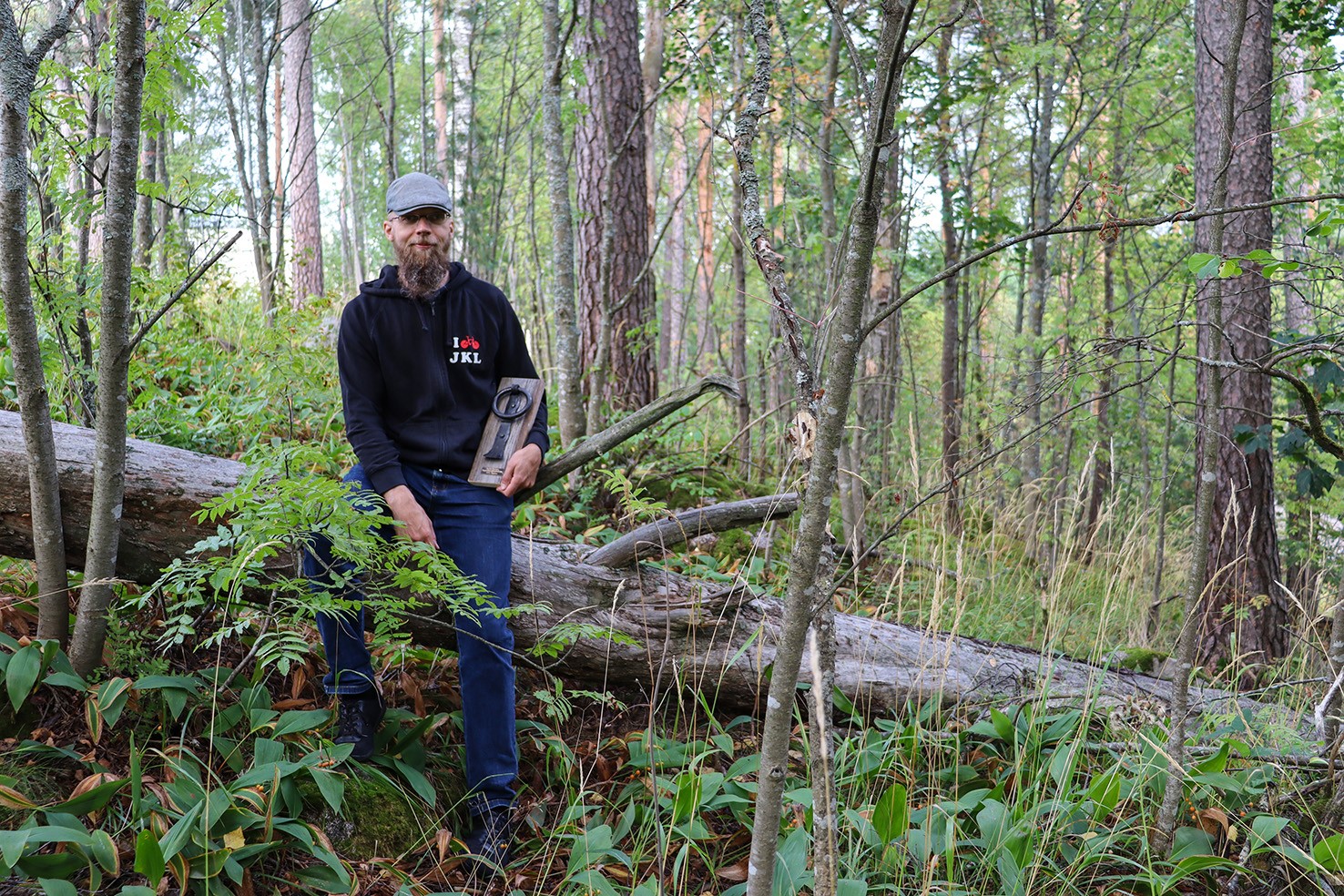 Juho Jäppinen istuu valoisassa sekametsässä kaatuneella puunrungolla ja pitää kädessään Kolkutin-veistosta.