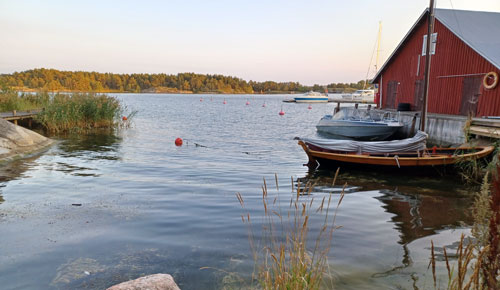 Tyyni Saaristomaisema. Kuvassa näkyy kaksi venettä, joista toinen on perinteinen puuvene ja toinen uudempi moottorivene. Veneiden takana näkyy punainen venevaja.